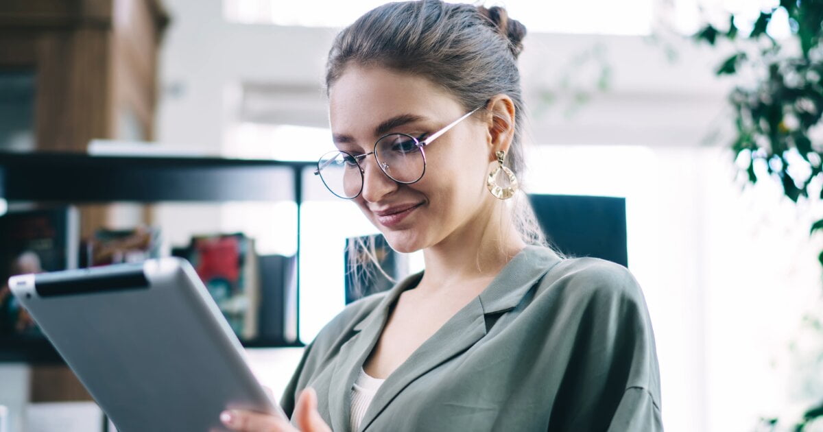 Bookstore image of a smiling young woman with brown hair, in a green jacket, as she is reading from her digital tablet.