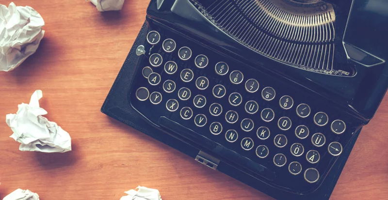 An old typewriter with the keyboard viewed from above on a wooden desk, where crumpled up sheets of paper are lying.