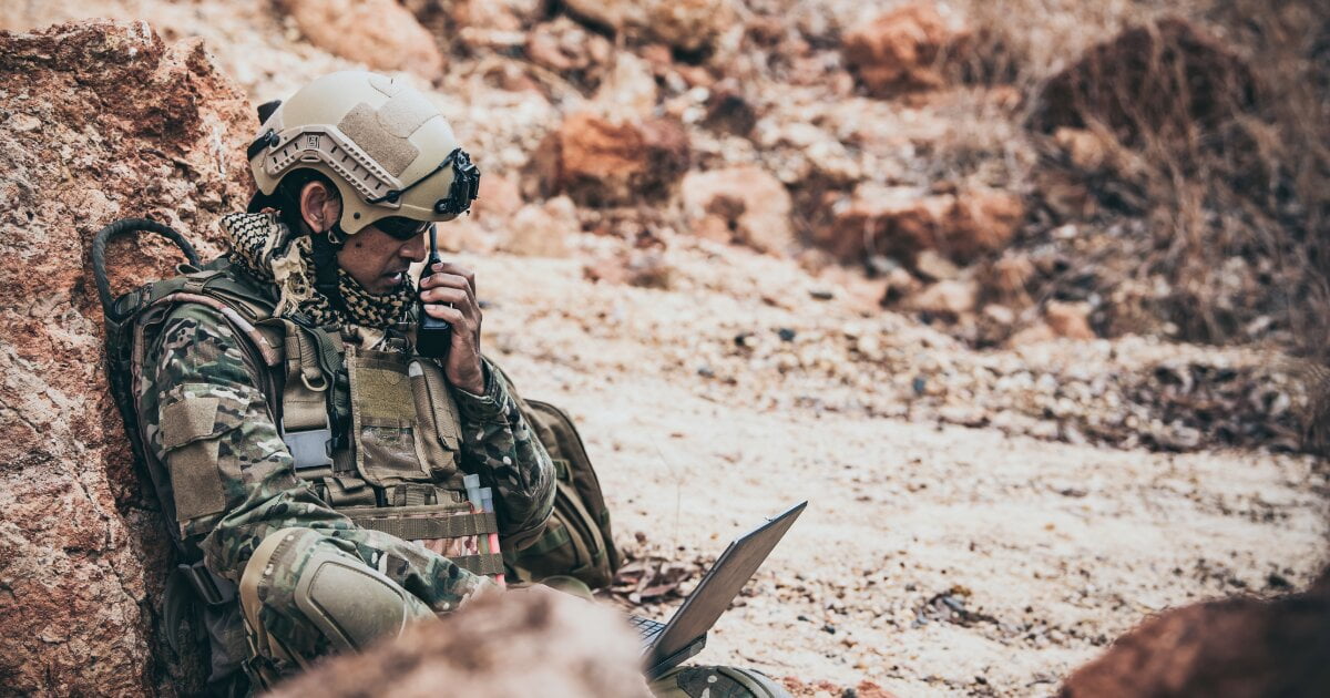 Special Operator lying in cover on the side of a dirt track calling in tactical coordinates using his walkie and his tactical laptop.