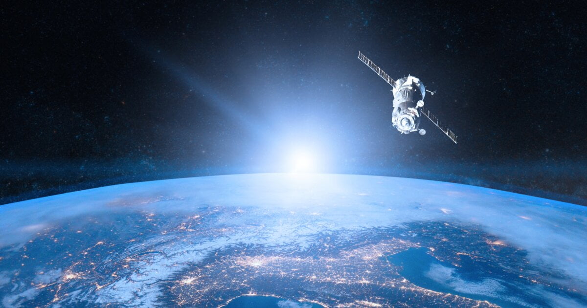 Space born view of near-earth with lit settlements below, and the rising sun in the background and a satellite to the upper right view.