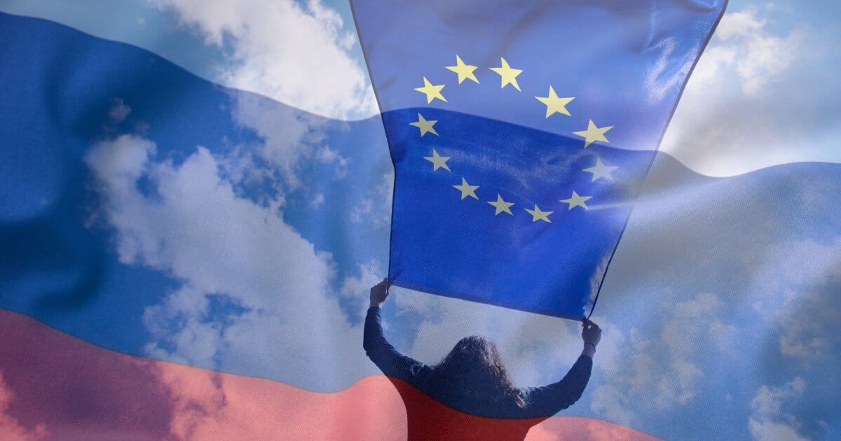 Woman holds up EU flag to the breeze with the blue sky overhead, but in the opague shadow of the Russian flag.